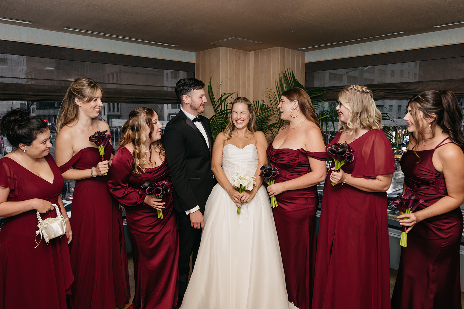 Wedding party portrait at Acadia NYC with the bride and groom surrounded by bridesmaids in deep red dresses and groomsmen in black tuxedos.