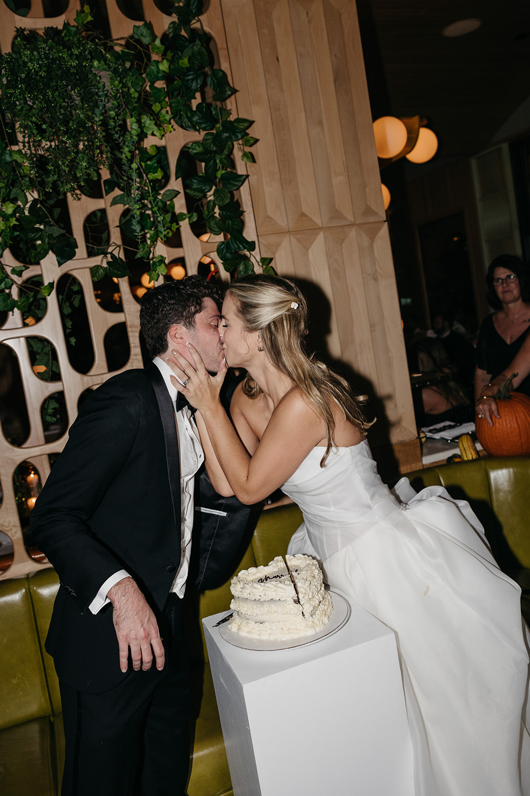Bride and groom sharing a kiss during cake cutting inside Acadia NYC with modern wood paneling and greenery behind them.
