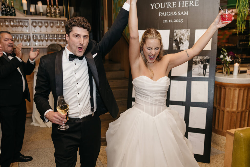 Bride and groom celebrating with raised arms holding drinks in front of their seating chart at Acadia NYC.