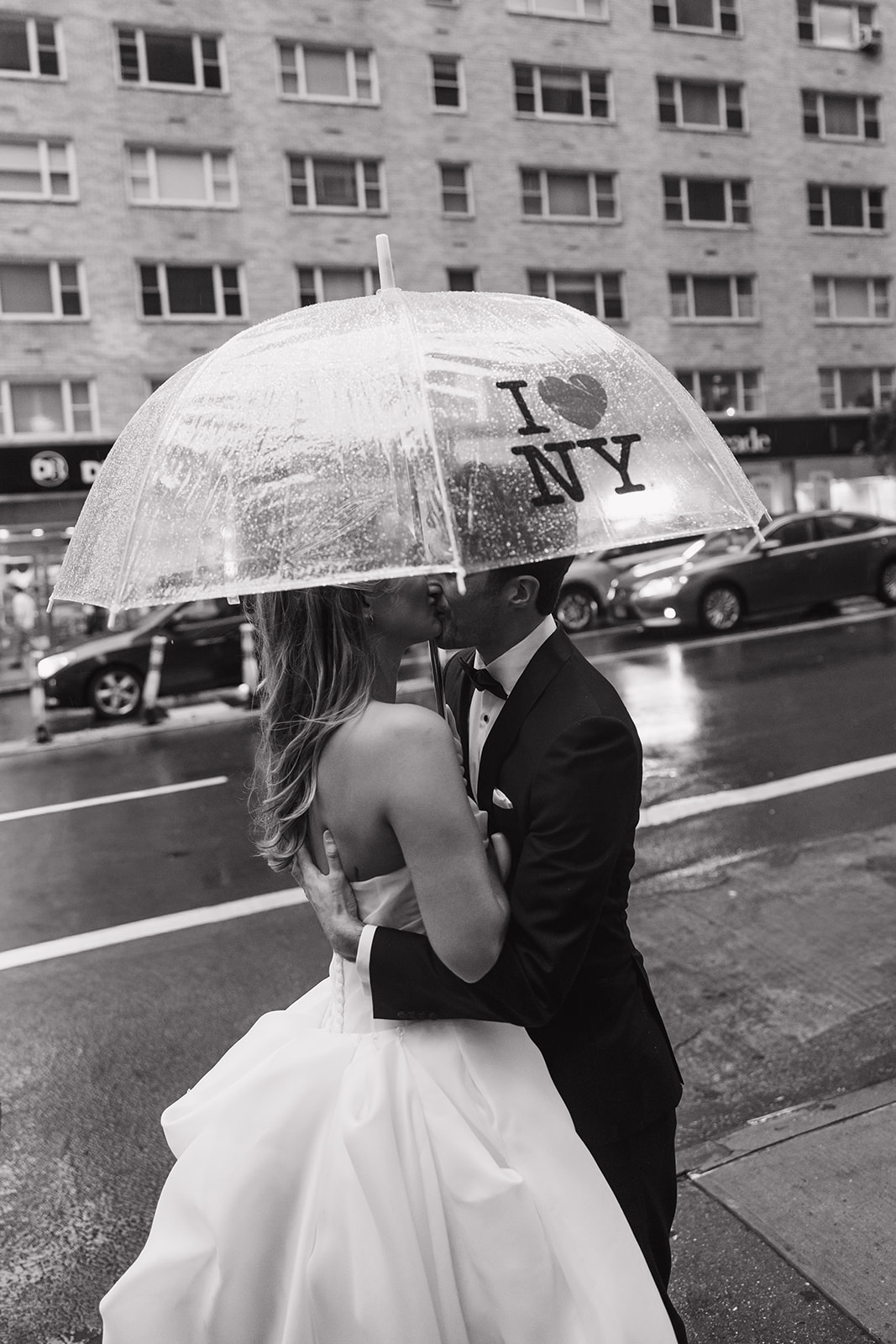 Black and white rainy NYC wedding photo of the couple kissing under an I Love NY umbrella on the sidewalk.