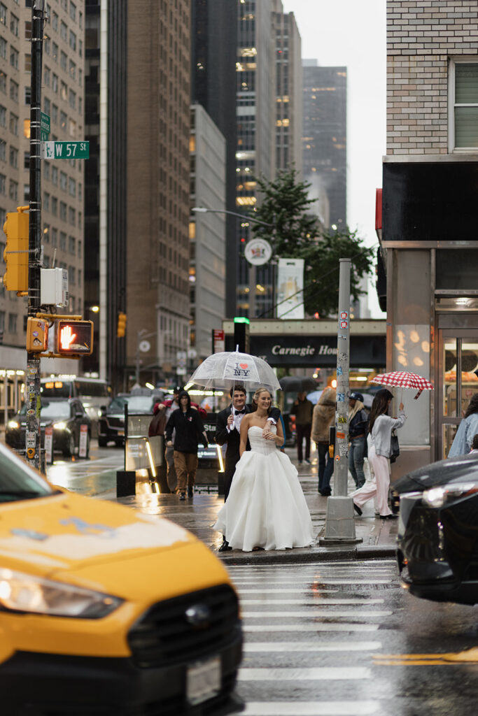 Bride and groom crossing a rainy Manhattan street holding umbrellas with taxis and city lights behind them.