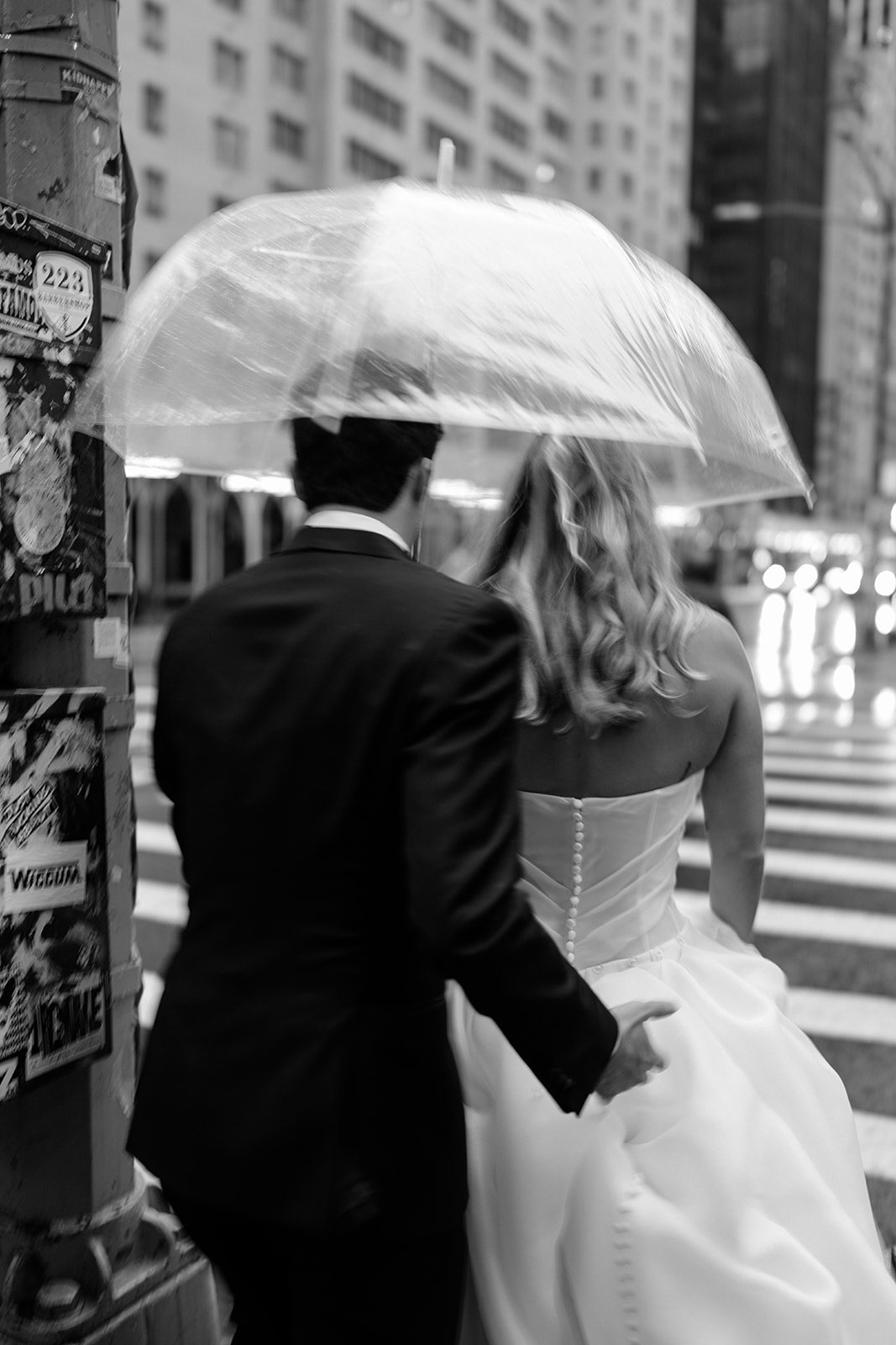 Black and white candid wedding photo from behind showing the couple walking together under a clear umbrella in NYC.