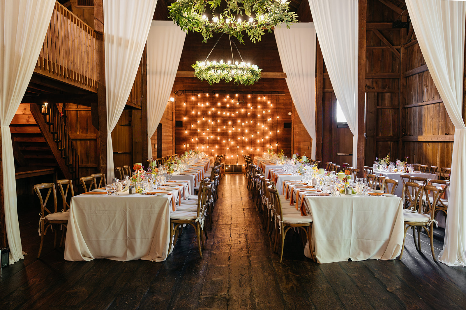 Wide view of the barn interior at The Barns at Wesleyan Hills CT wedding showcasing wooden beams, white drapery, greenery chandeliers, and warm lighting.