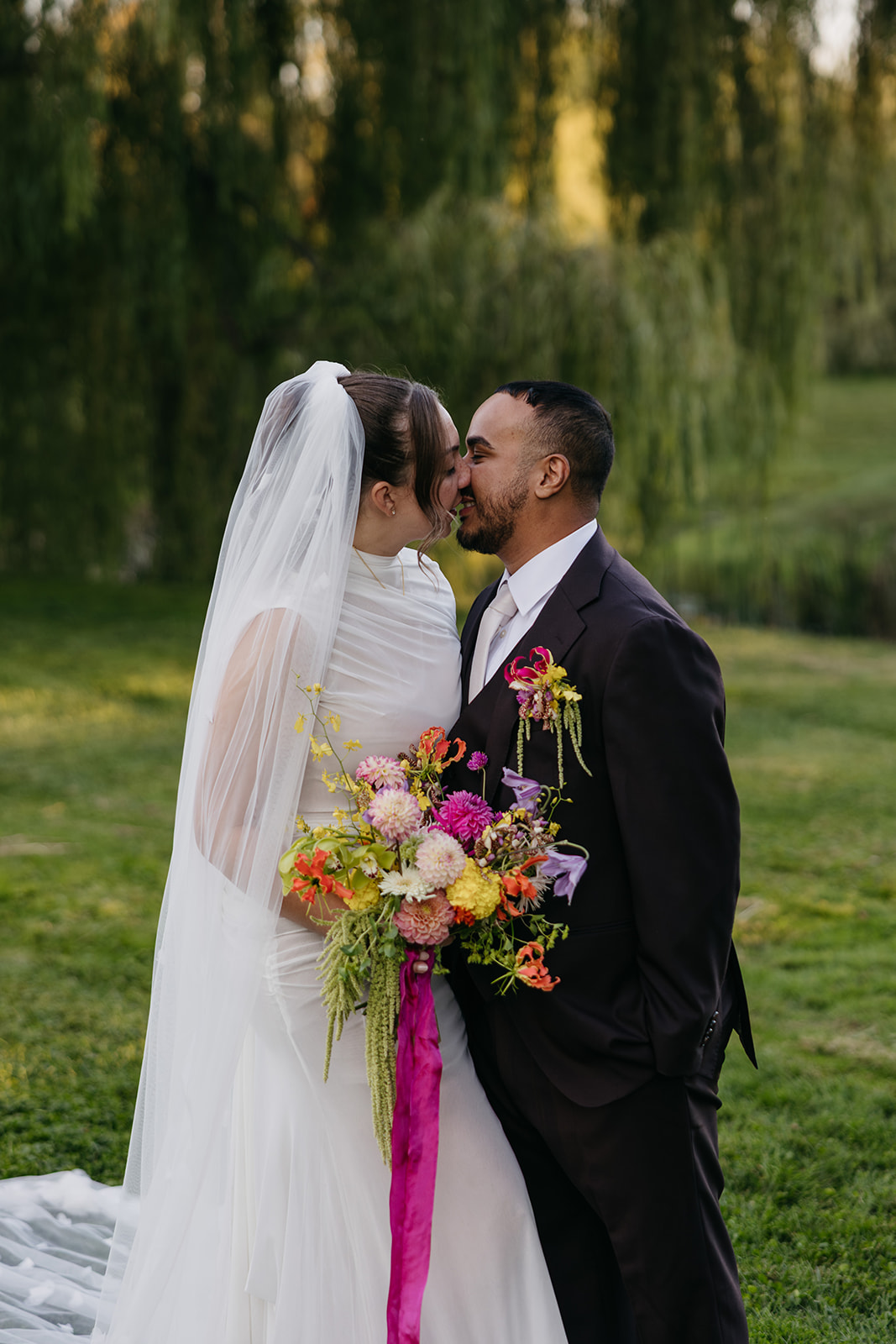 Bride and groom sharing a kiss outdoors at their Connecticut wedding surrounded by lush willow trees and colorful bouquet details.