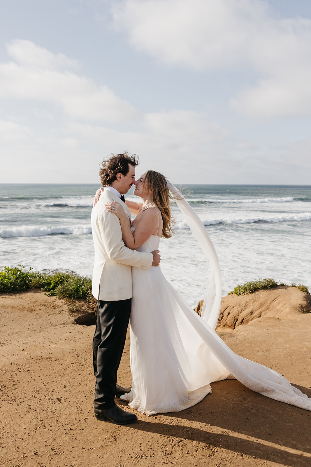Bride and groom holding each other by the ocean with veil flowing in the wind during their san diego wedding