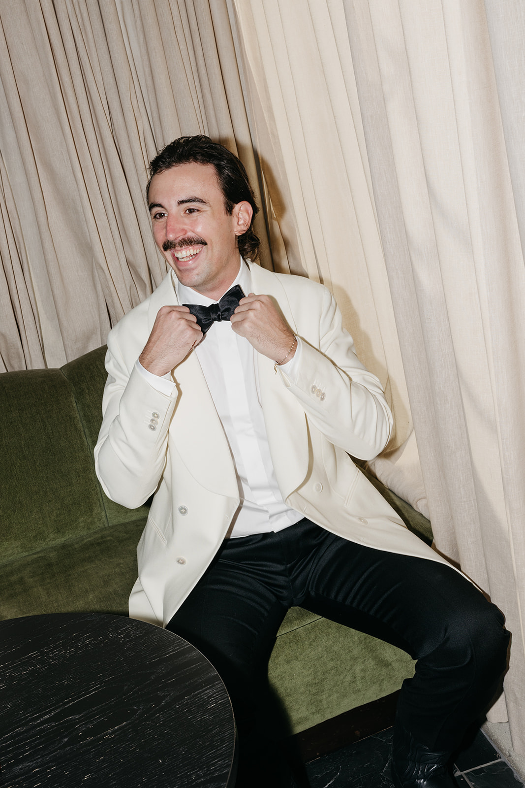 Groom adjusting bow tie while sitting in hotel room before ceremony